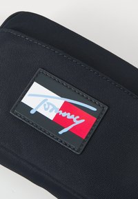 Granatowa torba materiałowa z prostokątną naszywką z logo w kolorze czerwono-białym i niebieskim ręcznie pisanym napisem "Tommy". Teksturowana powierzchnia.