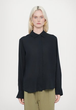 Schwarze Bluse mit langen Ärmeln, spitzem Kragen, Knopfleiste und weicher Textur; kombiniert mit hellgrünen Jogginghosen mit Kordelzug.