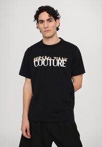 Juodas medvilninis marškinėliai su apvalia kaklo iškirpte, ant priekio esantis baltas ir bežinis tekstas "VERSACE JEANS COUTURE".