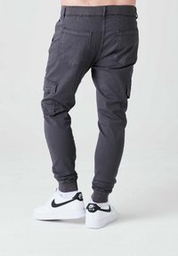 Šedé cargo kalhoty mají zúžený střih s elastickými manžetami a několika kapsami; nošeny s bílými teniskami s černými akcenty.