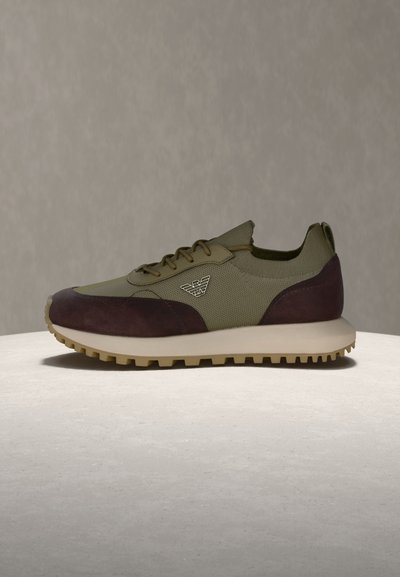 Ανδρικό αθλητικό παπούτσι σε ελιά πράσινο και σκούρο καφέ χρώμα με υφασμένο πέλμα και έμβλημα λογότυπου στην πλευρά, εκτεθειμένο σε στρογγυλή μπεζ επιφάνεια.
