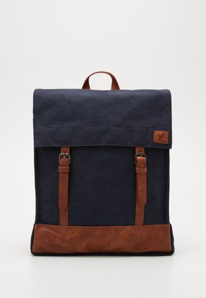 Sac à dos rectangulaire bleu marine avec des bretelles en cuir marron, un bord inférieur, une poignée supérieure et un petit patch en cuir sur le rabat.