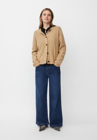 Beige gestrickte Strickjacke mit schwarzen Knöpfen, kombiniert mit weit geschnittenen dunkelblauen Jeans und gemusterten spitzen Schuhen.
