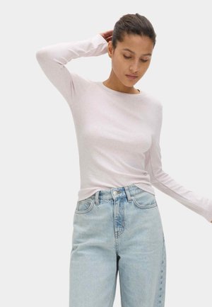 Mujer que lleva una camisa de manga larga de color rosa claro y unos vaqueros de tiro alto azul claro, con una mano detrás de la cabeza y mirando hacia abajo.