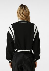 Černá zkrácená bunda s bílými pruhovanými akcenty, žebrovanými manžetami a límcem. Materiál se jeví hladký. Design zahrnuje detail loga na rukávu.