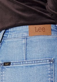 Světle modré džíny z denimu s hnědou koženou nášivkou s logem "Lee" na pase a malou černou etiketou na bočním švu.