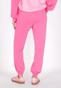 Rózsaszín adidas nadrág, amely puha anyagból készült, rugalmas mandzsettákkal, laza szabással és minimális varrási részletekkel rendelkezik. Világos rózsaszín sportcipővel párosítva.