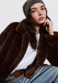 Manteau en fausse fourrure marron avec un grand col, porté sur une chemise blanche et un bonnet en tricot gris. Pantalons en denim bleu décontractés avec une ceinture marron.