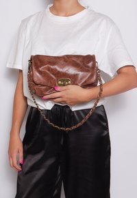 Borsa a pochette in pelle marrone con finitura lucida, hardware color oro, pattina pieghevole e tracolla a catena. Il modello la tiene contro uno sfondo bianco.