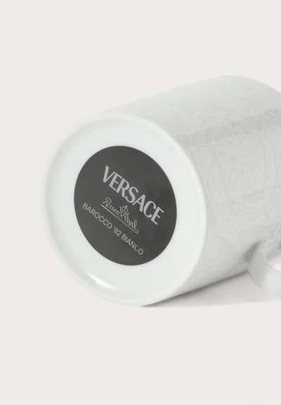 Tasse en céramique blanche Versace Barocco '92 Bianco avec des motifs floraux complexes, présentée couchée sur le côté mettant en évidence l’étiquette du dessous.
