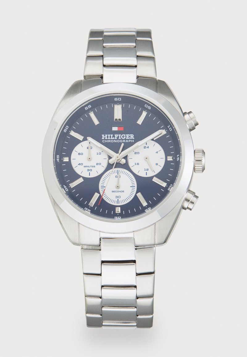 Ασημένιο ρολόι χρονογράφου με ναυτικό μπλε καντράν, λευκούς υποκαντράν και μπρασελέ από ανοξείδωτο ατσάλι. Περιλαμβάνει φωτεινές ενδείξεις ωρών και ένδειξη ημερομηνίας.