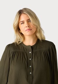 Blouse olive verte à manches longues avec un col rond, fermeture à boutons, détails plissés et un tissu doux et texturé.