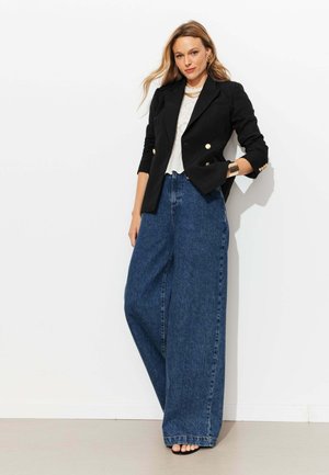 Femme aux longs cheveux blonds portant un blazer noir croisé, un top blanc, un jean bleu à jambes larges et des chaussures noires à bouts ouverts, debout contre un mur blanc.