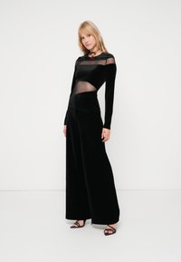 Fato macacão preto em veludo com mangas longas, detalhes em malha transparente, pernas largas e cintura ajustada. Combinado com sapatos de salto pretos com tiras.