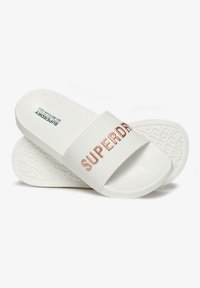 White slide sandals with a thick band featuring embossed "SUPERDRY" in rose gold. Smooth material, textured sole for grip.
