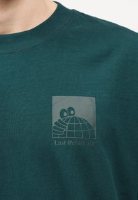 Σκούρο πράσινο πουκάμισο με ένα μικρό λογότυπο ενός καρτούν χεριού που κρατάει μια υδρόγειο και το κείμενο "Last Resort AB" στο στήθος.