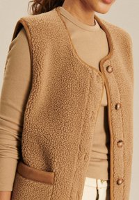 Beige Fleece-Weste mit strukturierter Oberfläche, rundem Ausschnitt, drei Knöpfen auf der Vorderseite und seitlichen Taschen. Über einem taillierten, beigen Oberteil gestylt.