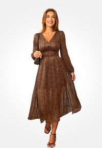 Brązowa maksi sukienka w kwiaty z długim rękawem, dekoltem w kształcie litery V i marszczoną talią. Lekka tkanina z lekko opadającą spódnicą i bocznymi rozcięciami. Czarne sandały.
