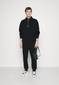 Sudadera negra con cremallera y logo bordado, combinada con joggers negros y zapatillas blancas con acentos en negro y rojo. Bolsa negra en la mano.
