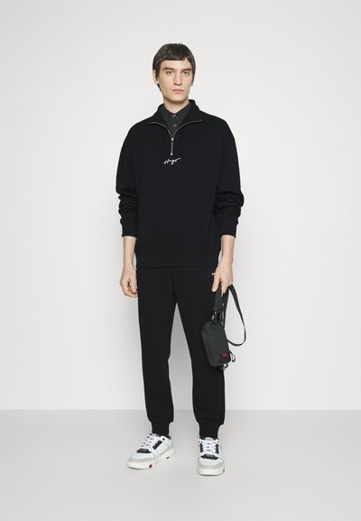 Sweatshirt zippé noir avec logo brodé, associé à un jogging noir et des baskets blanches avec des accents noirs et rouges. Sac noir à la main.