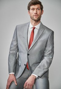 Világosszürke öltöny sima anyagból, amelynek felső része kiemelkedő hajtóka, két gomb és elülső zsebek találhatók. Piros nyakkendővel és fehér inggel párosítva.