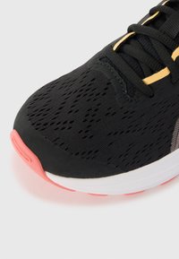 Chaussure de sport noire avec une tige texturée, dotée d'une semelle blanche et rose, et d'accents dorés sur le système de laçage.