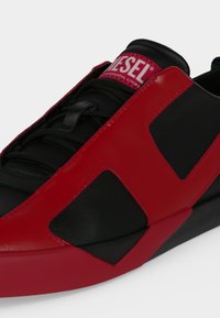 Baskets noires avec des surpiqûres rouges audacieuses et une étiquette rouge au logo Diesel sur la languette, montrant un gros plan de la tige et des lacets.