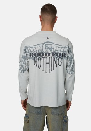 Chemise à manches longues gris clair avec un grand motif d'ailes et le texte « GOOD FOR NOTHING » en noir gras, avec une étoile au-dessus.