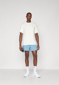 Balta T-kreka ar īsām piedurknēm un zili sporta šorti. Modelis tur ūdens pudeli. Balti sneki ar melniem akcentiem. Vienkārša fona.