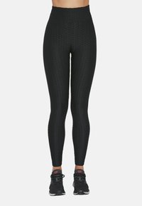 Czarne legginsy z wysokim stanem wykonane z fakturowanej tkaniny o wzorze plastra miodu, zestawione z czarnymi butami sportowymi z różowymi akcentami.