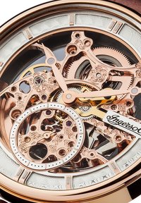Orologio scheletrato con ingranaggi intricati in tonalità oro rosa, movimento visibile e un quadrante bianco texturizzato. Presenta un piccolo sottodial per i secondi e un accento con marchio.