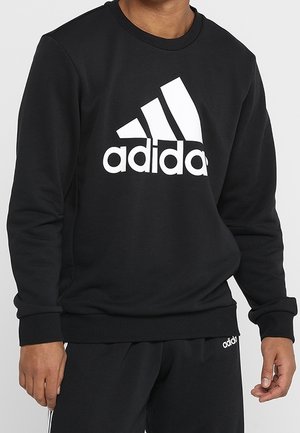 Mężczyzna w czarnej bluzie Adidas z dużym białym logo na klatce piersiowej oraz czarnych spodniach z małym logo Adidas.