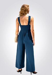 Navyblauer Jumpsuit mit weiten Beinen, gerafftem Oberteil und dicken Trägern. Das Model hält eine gewebte Clutch und trägt offene Schuhe mit Absatz.