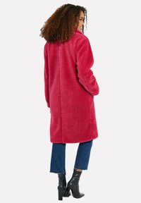 Lång, mysig, rosa skinnjacka i fårskinn med hög krage och två sidofickor, tillsammans med mörka korta jeans och ankelstövlar.