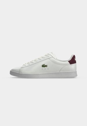 Baskets basses blanches avec lacets blancs, logo crocodile vert sur le côté et languette de talon rouge foncé sur semelle grise.