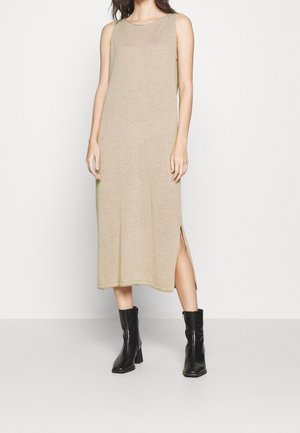Mouwloze beige midijurk met een zijsplit, gedragen met zwarte enkellaarzen met hak, staand tegen een witte achtergrond.