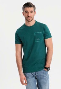 Teal bomullströja med rund hals och bröstficka, med en fyrkantig logotyp på fickan. Modellen bär den med ljusblå jeans.