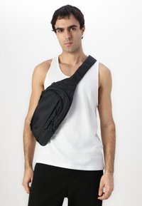 Sac bandoulière noir en nylon, avec une texture lisse, plusieurs poches zippées et une large sangle pour un port en travers.