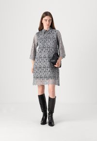 Robe en dentelle grise avec un col, des manches trois-quarts et un ourlet festonné. Portée avec des bottes noires à hauteur de genoux et un petit sac.