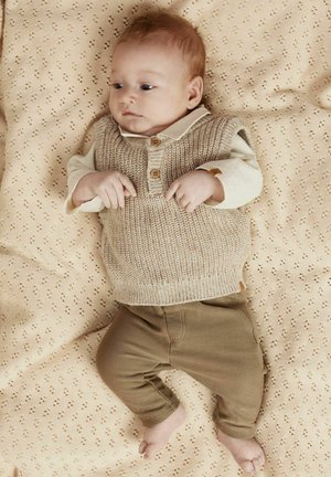Gilet lavorato a maglia di un beige chiaro con bottoni in legno sopra una camicia crema e pantaloni verde oliva, adagiato su una coperta beige a fantasia.