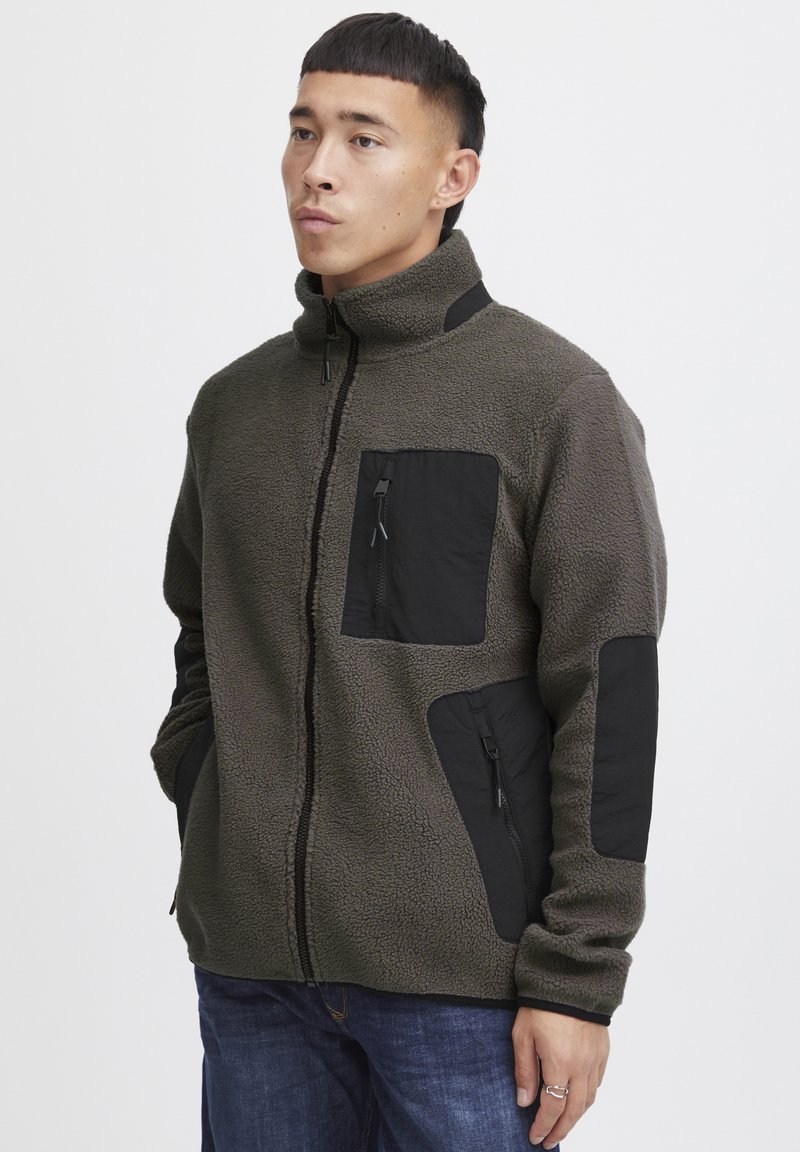 Fleece jack in olijfgroen met zwarte nylon accenten. Voorzien van een hoge kraag, een ritssluiting aan de voorkant en meerdere ritszakken.