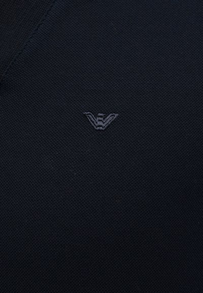 Σκούρο υφασμάτινο με υφή, με ένα μικρό, κεντημένο γκρι λογότυπο που απεικονίζει ένα στυλιζαρισμένο «W» στο κέντρο.