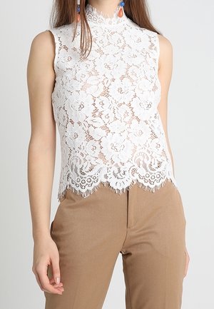 Ärmelloses weißes Spitzenoberteil mit Blumenmuster, getragen zu hochgeschnittener beiger Hose, sichtbare blaue und orangefarbene Tropfenohrringe.