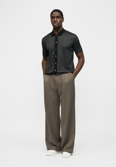 Άνδρας που φοράει μαύρο πουκάμισο με μοτίβα και κοντά μανίκια με κουμπιά, μπεζ παντελόνι φαρδιάς γραμμής, λευκά αθλητικά παπούτσια, στέκεται με τα χέρια στις τσέπες.