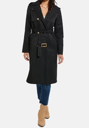 Femme portant un trench-coat noir ceinturé jusqu'aux genoux, un jean bleu raccourci et des bottines en daim couleur tan, debout devant un fond uni.
