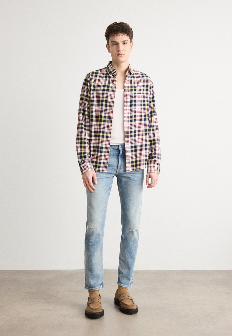 Camicia di cotone a quadri in rosso, blu navy e giallo; maniche lunghe, colletto alla francese. Indossata con jeans chiari strappati e scarpe slip-on marroni.