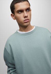 Sudadera verde claro con cuello redondo, hombros caídos y un ajuste relajado. Textura suave, con una capa interior blanca en el cuello.