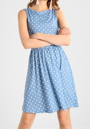 Femme portant une robe bleue sans manches à pois blancs et des boucles d’oreilles à pompons roses, bras croisés, sur un fond clair uni.