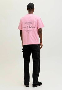 Kurzärmeliges pinkfarbenes Baumwoll-T-Shirt mit schwarzen Textgrafiken auf der Rückseite; kombiniert mit schwarzen Cargohosen und schwarzen Schuhen.