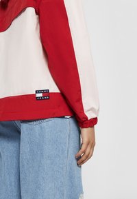 Czerwona i kremowa wiatrówka z elastycznymi mankietami i dołem, z widoczną metką z logo "TOMMY JEANS" na dolnym lewym boku. Pod spodem widoczne szorty jeansowe.